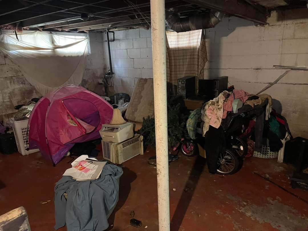 Cluttered basement before cleanout by Wilk's Junk Service, featuring a pink camping tent, scattered clothing, boxes, a bicycle, and a cat carrier, with dim lighting and cinder block walls.