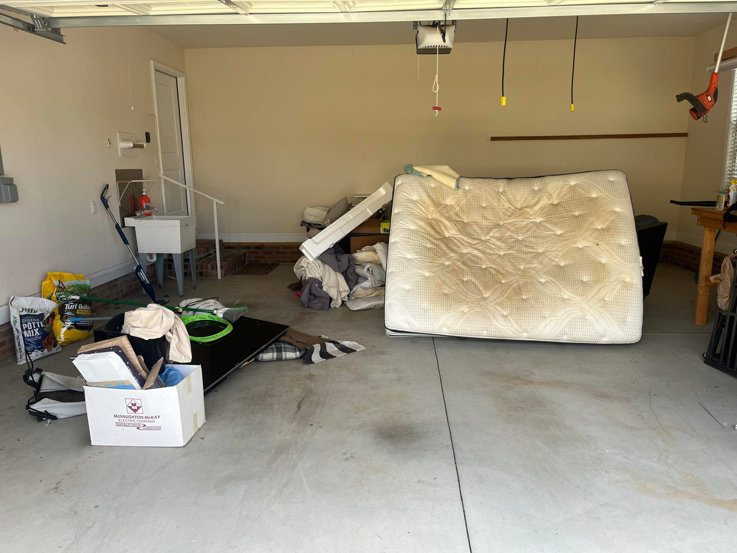 Garage cluttered with a mattress, bags, boxes, a bicycle, and tools before cleanout by Wilk's Junk Service, with natural light streaming in through the open garage door.