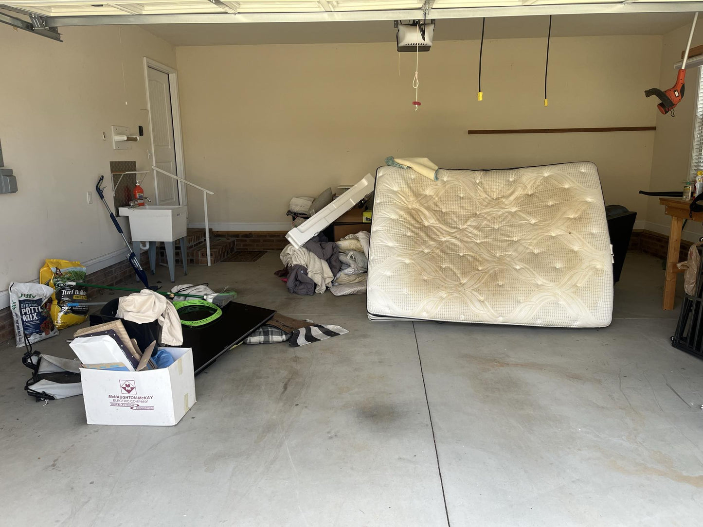 Garage cluttered with a mattress, bags, boxes, a bicycle, and tools before cleanout by Wilk's Junk Service, with natural light streaming in through the open garage door.