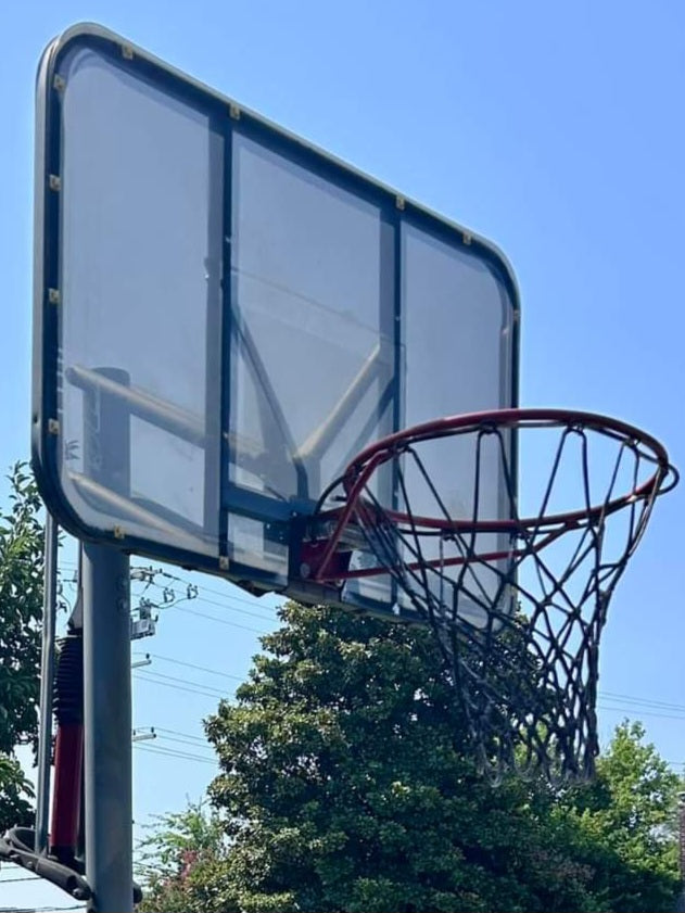 A basketball hoop with a clear backboard, attached to a red pole, with a bright blue sky and green tree leaves in the background. Removed by Wilk's Junk Service.