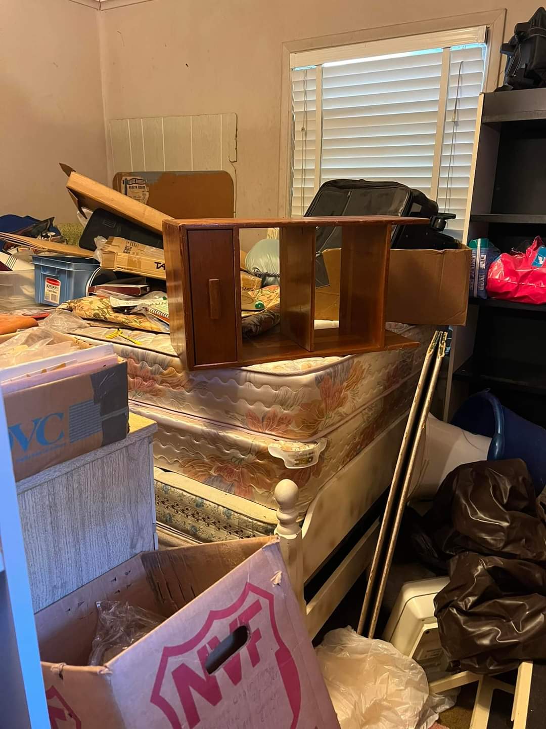 A cluttered room indicating a residential hoarding situation, with a mattress piled with miscellaneous objects, plastic bags, and boxes. This image represents a before-cleanup scenario for Wilk's Junk Service specializing in residential hoarding clean-up.