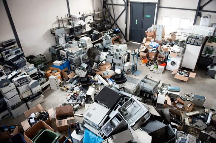 A cluttered warehouse filled with discarded electronic equipment, including computers, printers, and monitors. This image represents a warehouse cleanout scenario for Wilk's Junk Service.