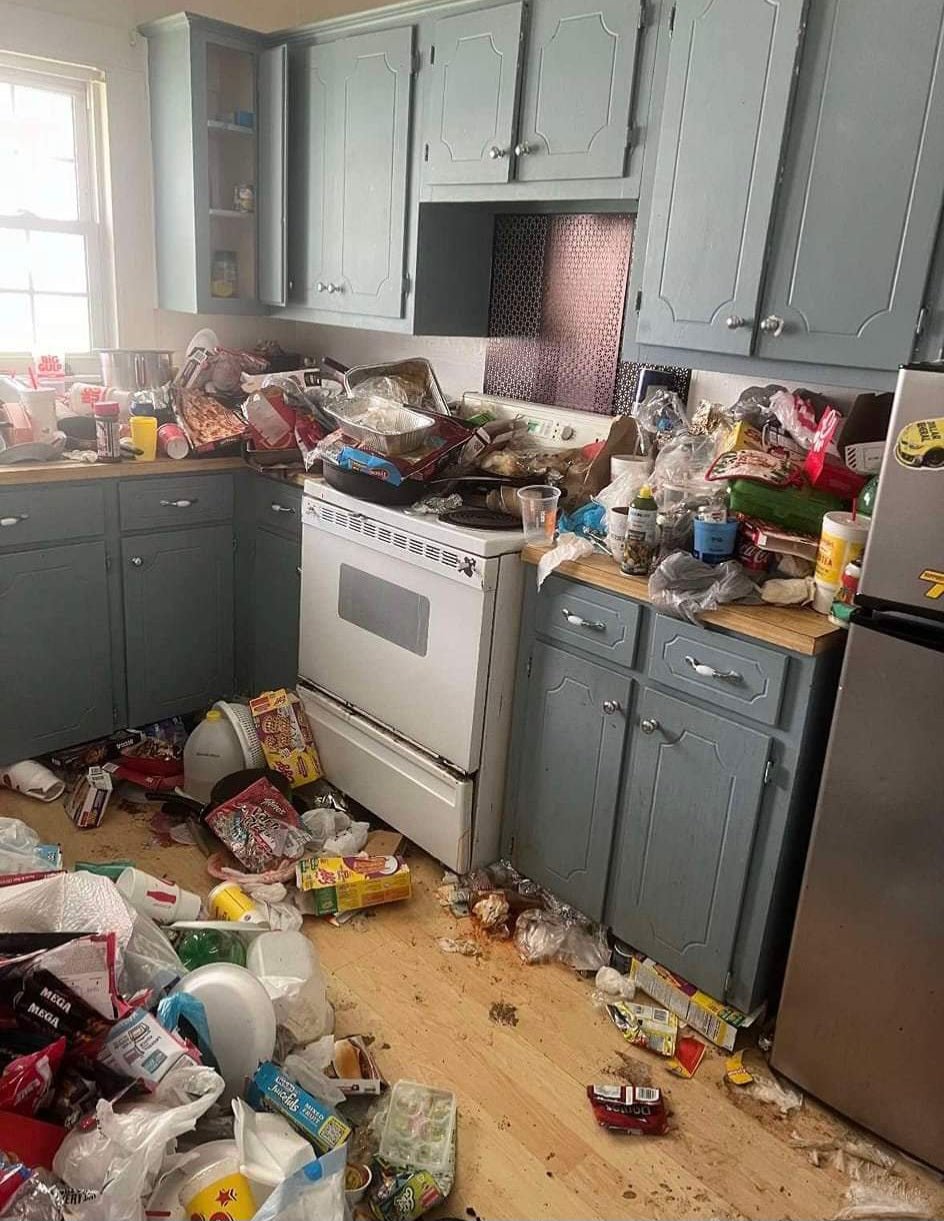 A kitchen in disarray with grey cabinets, a white stove, and a microwave. The floor and countertops are cluttered with food waste and various items, showing the state before Wilk's Junk Service.