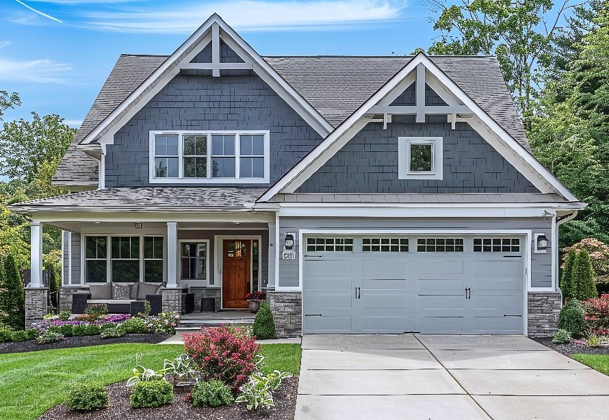 Two-story house with dark gray exterior, white trim, and a gabled roof. Covered front porch, two-car garage, and neat landscaping with hedges and flowers. Partly cloudy sky. Wilk's Junk Service.