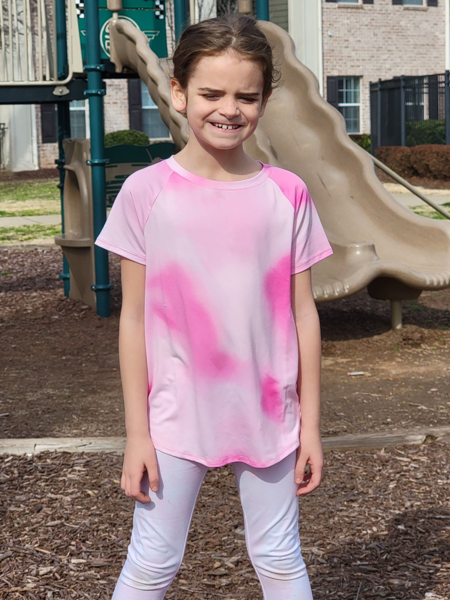 Bri standing outside in front of a playground slide wearing a pink shirt and white pants