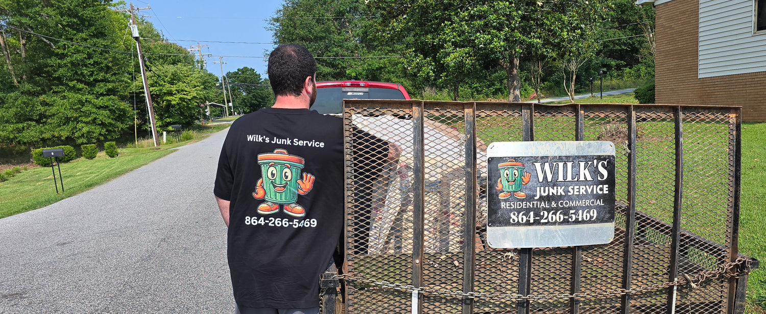 Brandon Costanza of Wilk's Junk Service stands beside a loaded trailer with the company logo and phone number (864-266-5469) in Spartanburg, SC, parked on a quiet residential street.